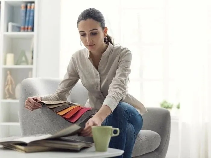 A woman seated in a grey armchair browsing colourful fabric samples, with a sample book and mug on a table