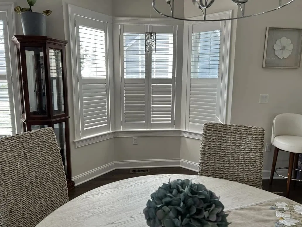 White cafe-style plantation shutters on a dining room bay window, viewed past woven chairs and a round table