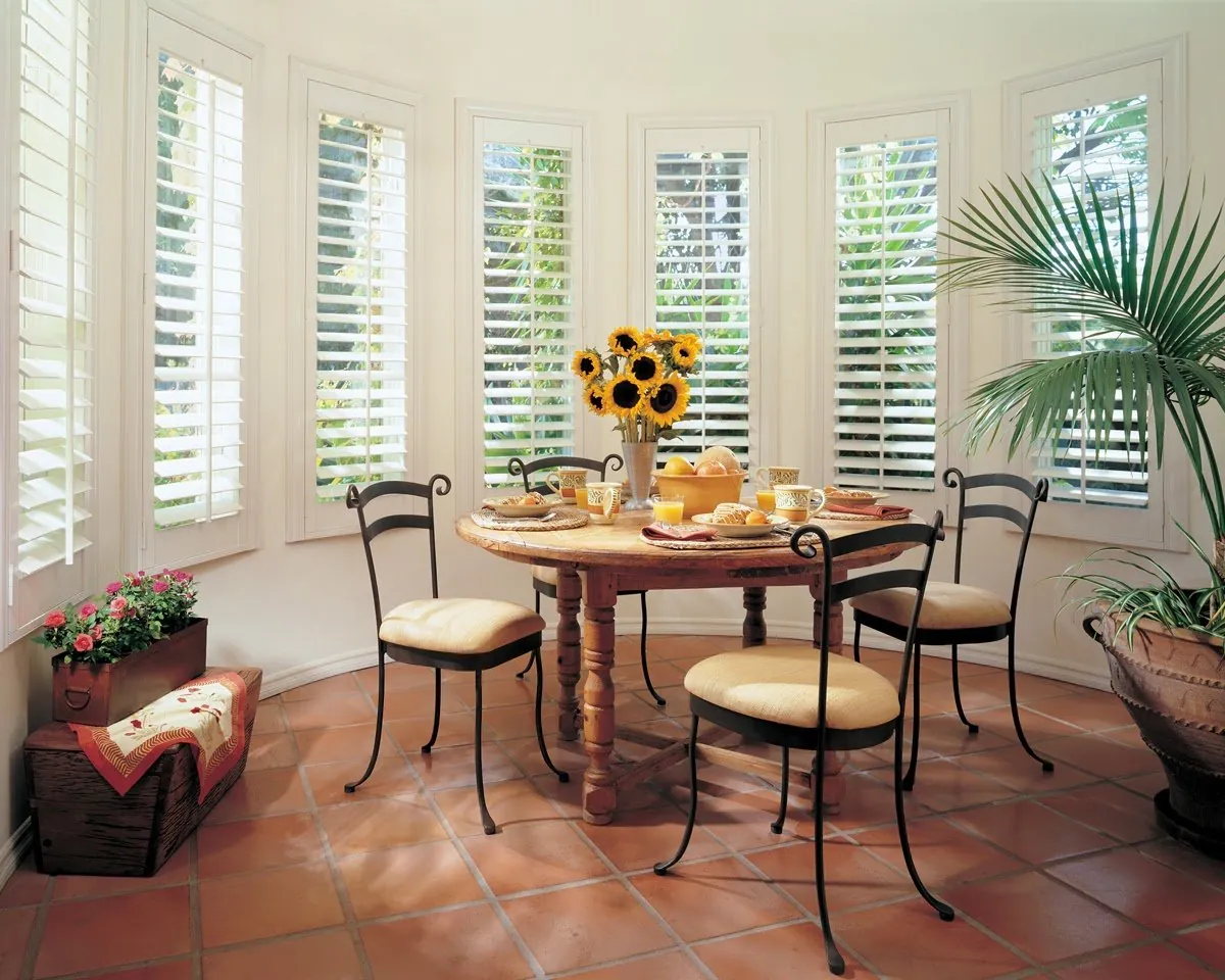 White plantation shutters on every window of a curved breakfast bay with a terracotta tile floor, a round table and sunflowers