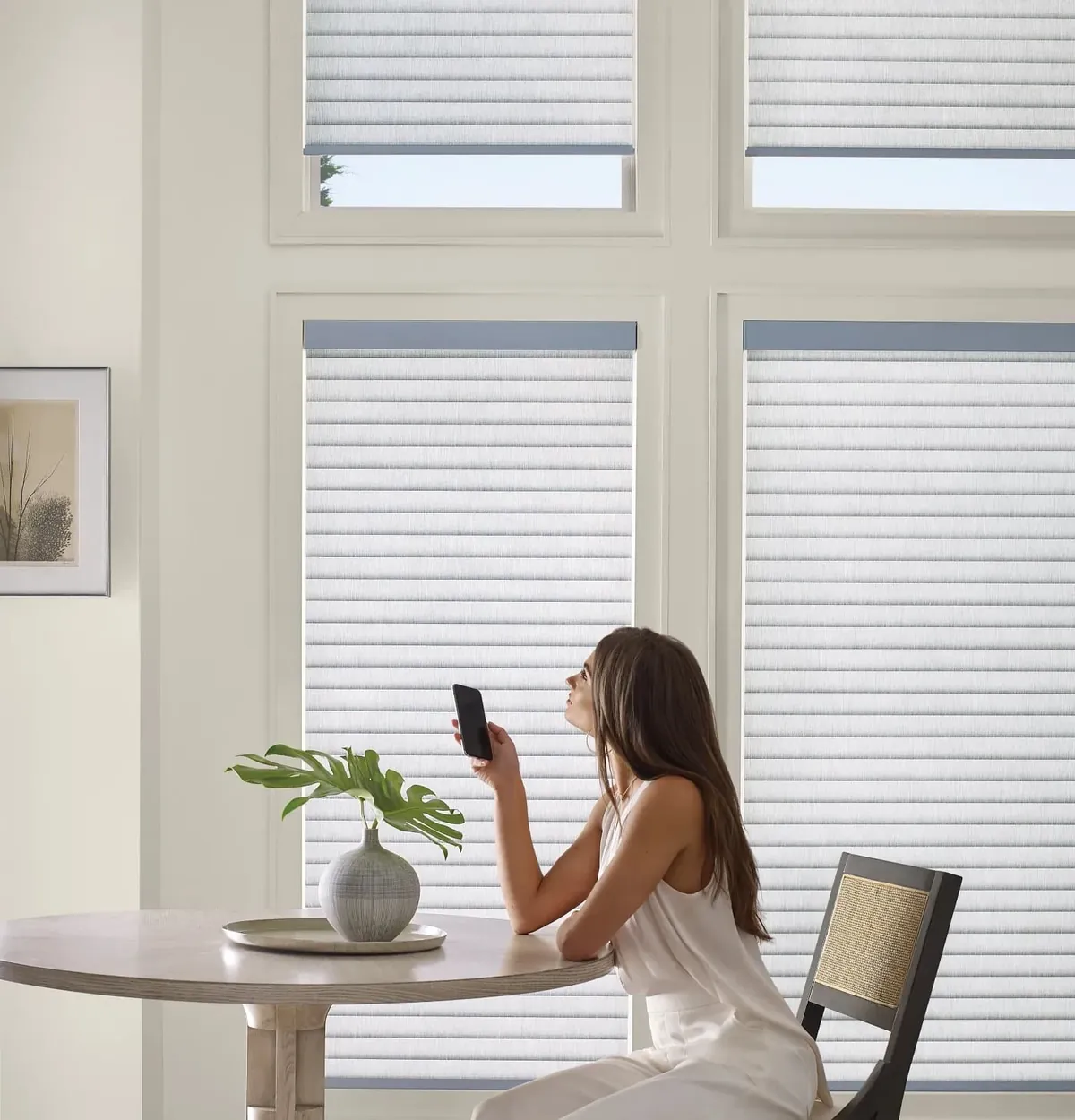 A woman at a round table holds up a phone to adjust soft white horizontal shades on tall windows and transoms