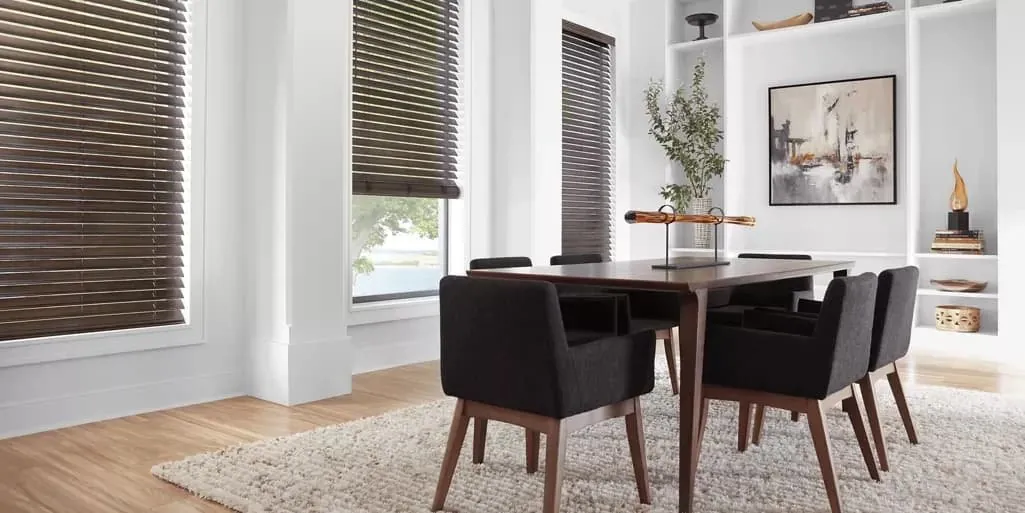 Dark brown wood blinds on tall windows in a white dining room with a wooden table and charcoal upholstered chairs