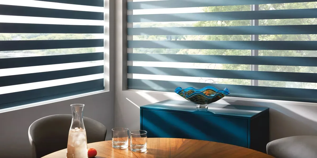 Deep teal banded shades with alternating sheer and solid stripes on corner windows above a round wooden table and blue cabinet
