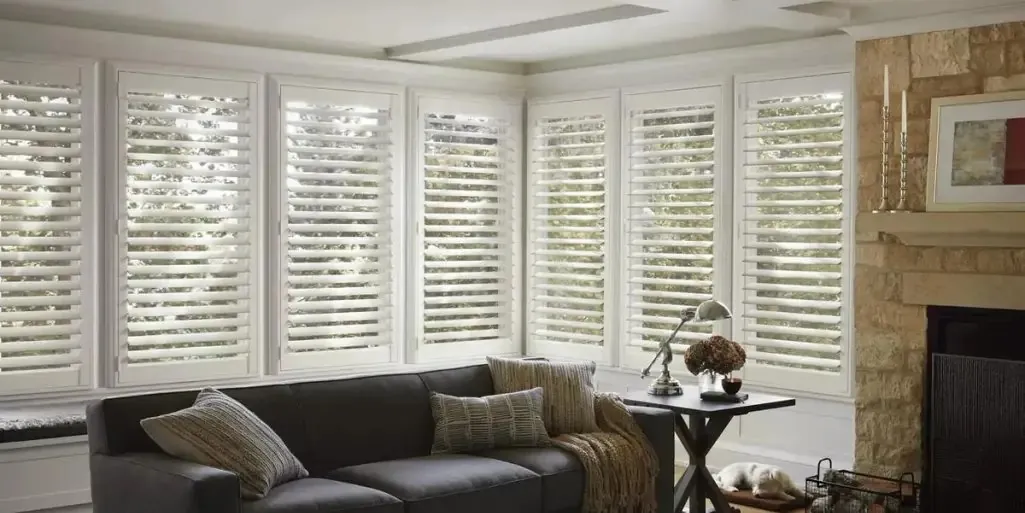 White shutters on a bay of windows behind a dark grey sofa, beside a stone fireplace, a dog asleep on the floor