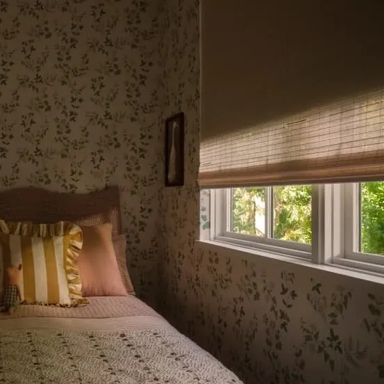 Natural woven shade on a bedroom window beside a bed with floral wallpaper and a striped ruffled pillow