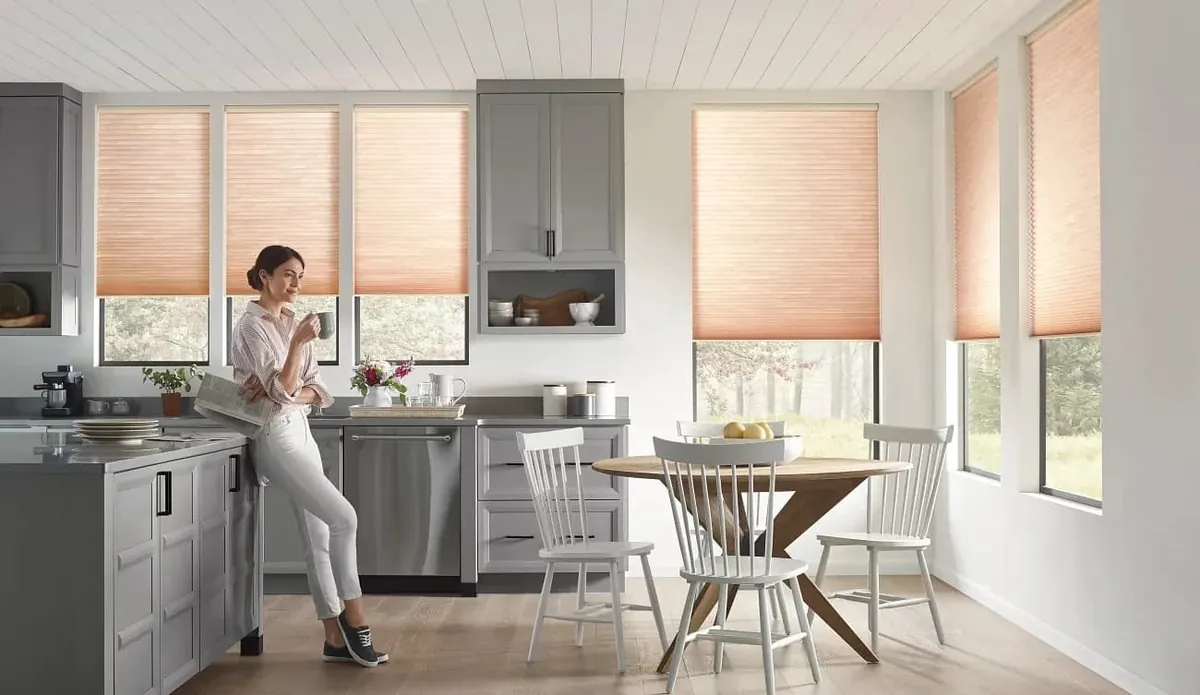 Peach light-filtering honeycomb shades half-raised on kitchen windows, with a woman drinking coffee by the island and a round dining table