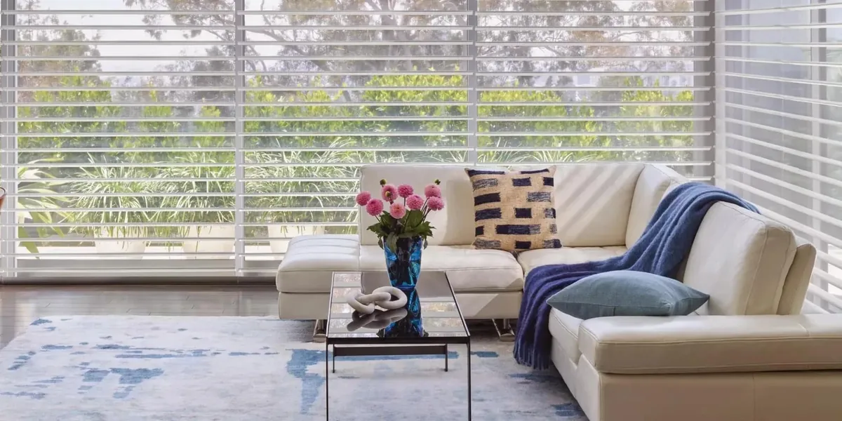 White sheer shades with horizontal vanes behind a cream leather sectional, glass coffee table and pink flowers, with garden greenery outside