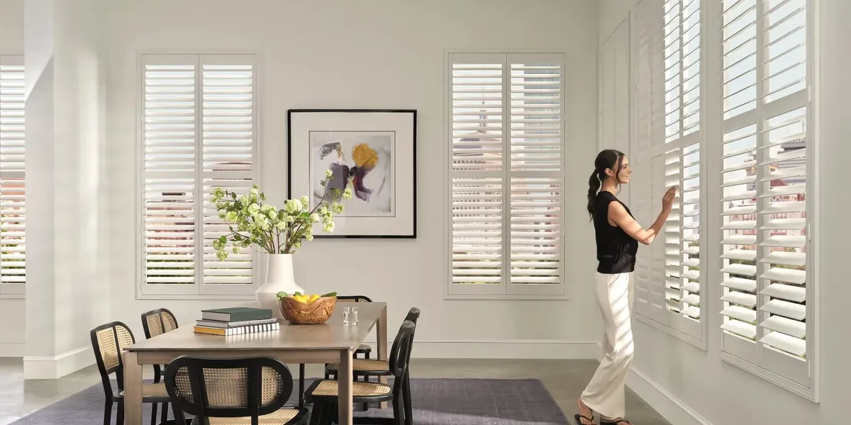 White plantation shutters on tall windows in a bright dining room, with a woman adjusting the louvres beside the table