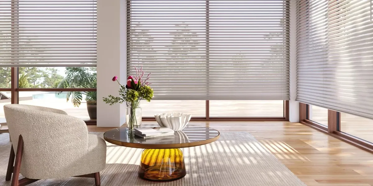 Light grey horizontal blinds partly raised on corner windows in a sunny lounge with a boucle chair and glass coffee table