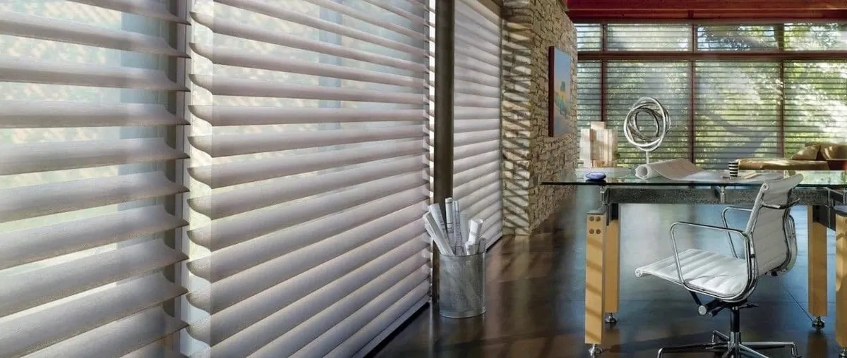 Grey sheer shades with floating fabric vanes along office windows, with a glass desk, white chair and stone wall