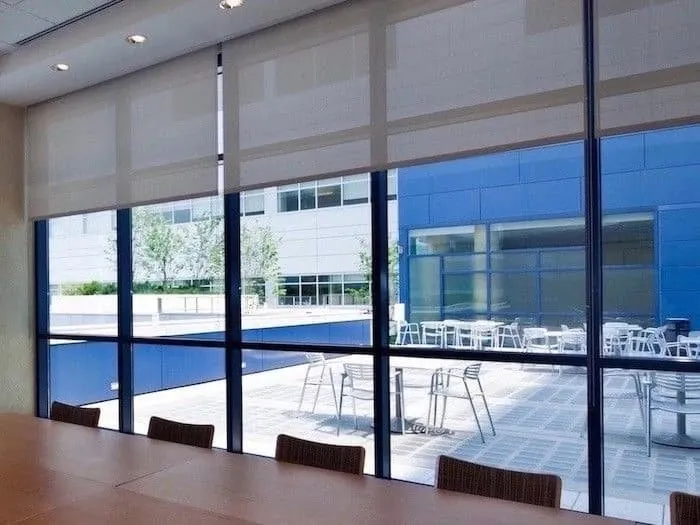 Light screen roller shades partly lowered on floor-to-ceiling windows of a conference room overlooking a courtyard patio