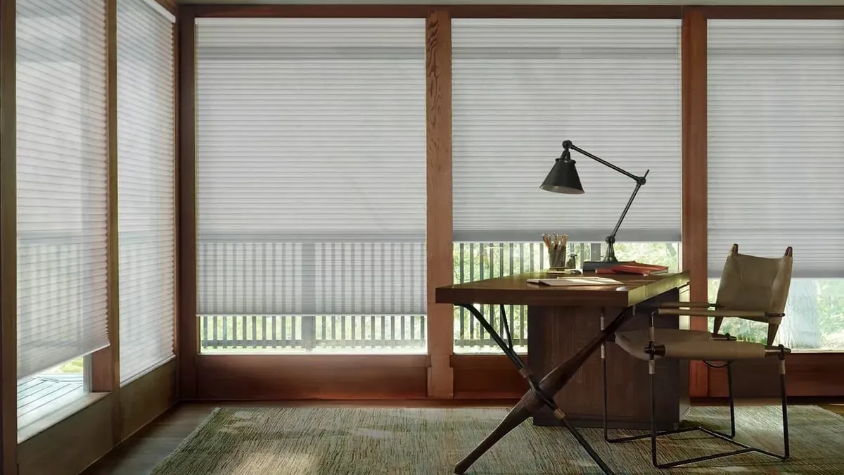 White honeycomb shades partly lowered on wood-framed windows in a home office with a desk, lamp and leather chair