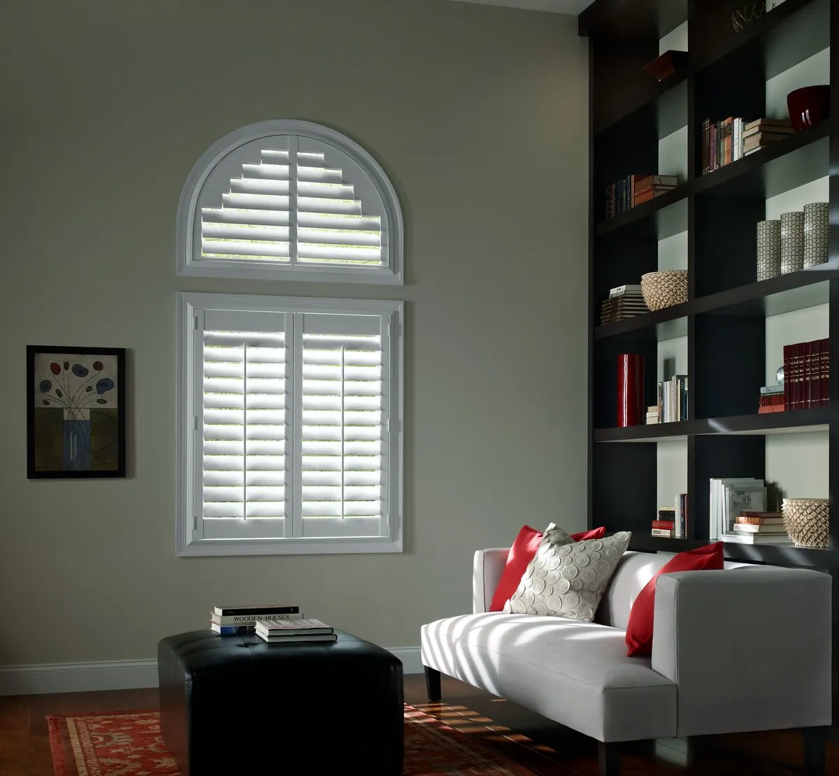 White plantation shutters on a tall window with a matching arched shutter above, beside dark built-in bookshelves and a white loveseat