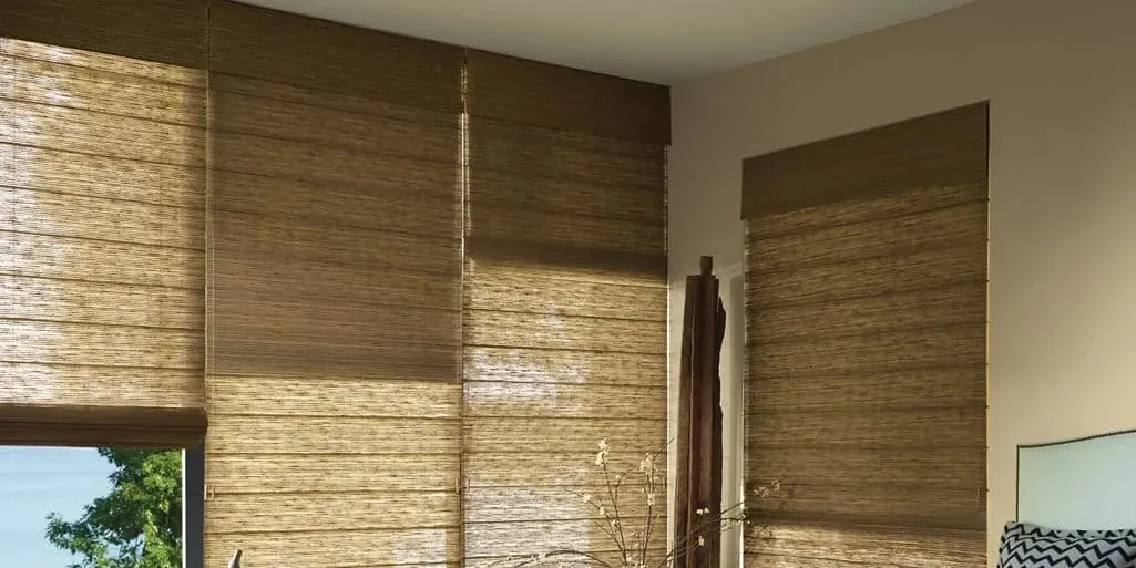 Natural woven roman shades on tall corner windows in a sunlit room, partly lowered with a view of trees