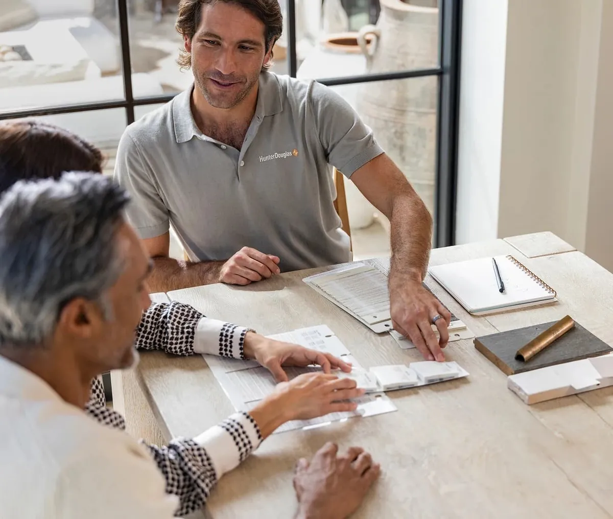 A consultant in a grey polo reviews shade sample books with a couple seated at a light wood table