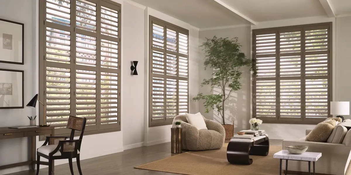 Taupe painted plantation shutters on tall living room windows, with a boucle armchair, sofa, potted tree and writing desk