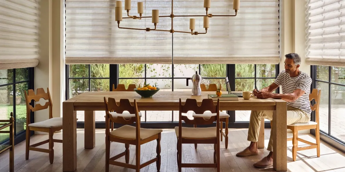 Light natural-weave soft-fold shades on large dining room windows, a man writing at a long wooden table