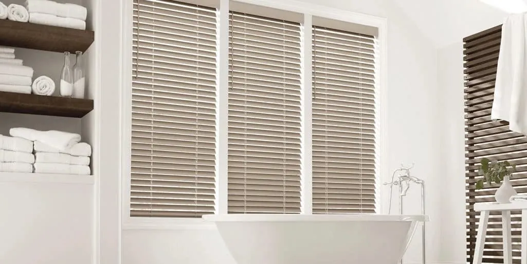 Taupe faux wood blinds on three windows above a freestanding white bathtub, with open shelves of folded white towels