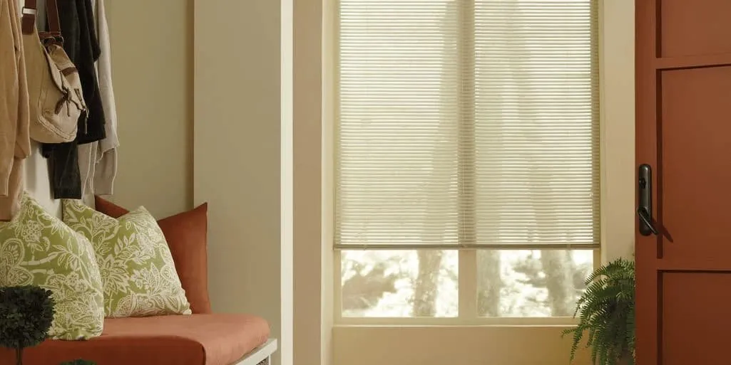 Cream metal mini blinds on an entryway window beside an orange door, a bench with patterned cushions and hanging coats