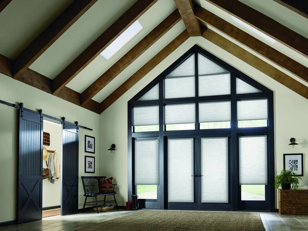White honeycomb shades on every pane of a tall gable window wall and French doors in a vaulted room with wood beams and a black barn door