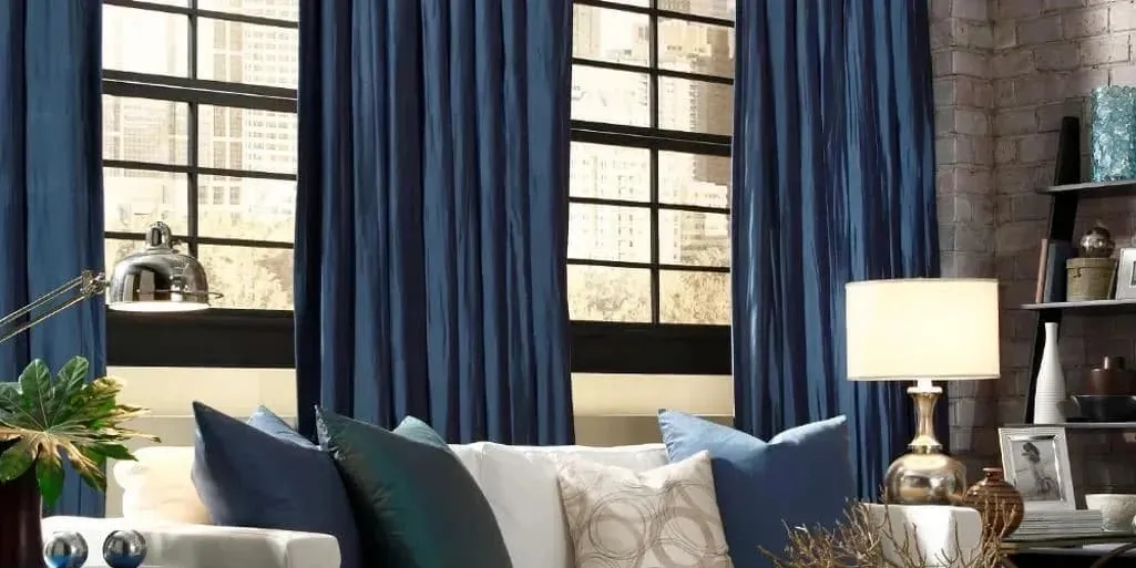 Navy blue drapery panels at tall black-framed loft windows behind a white sofa with blue cushions and a brick wall