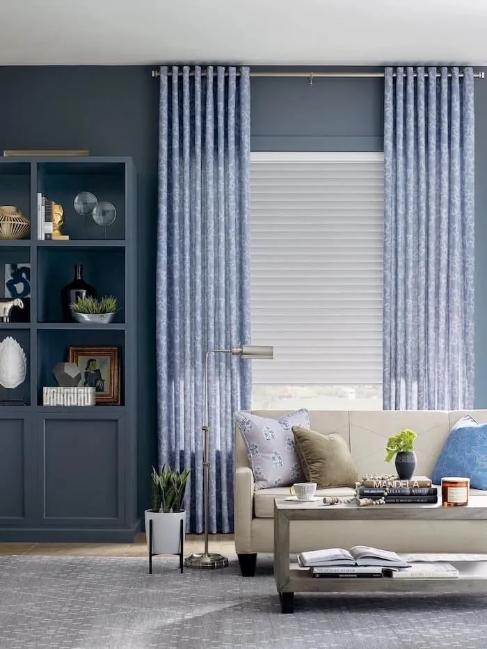 Blue patterned drapery panels framing a white sheer shade behind a cream sofa in a navy-walled living room