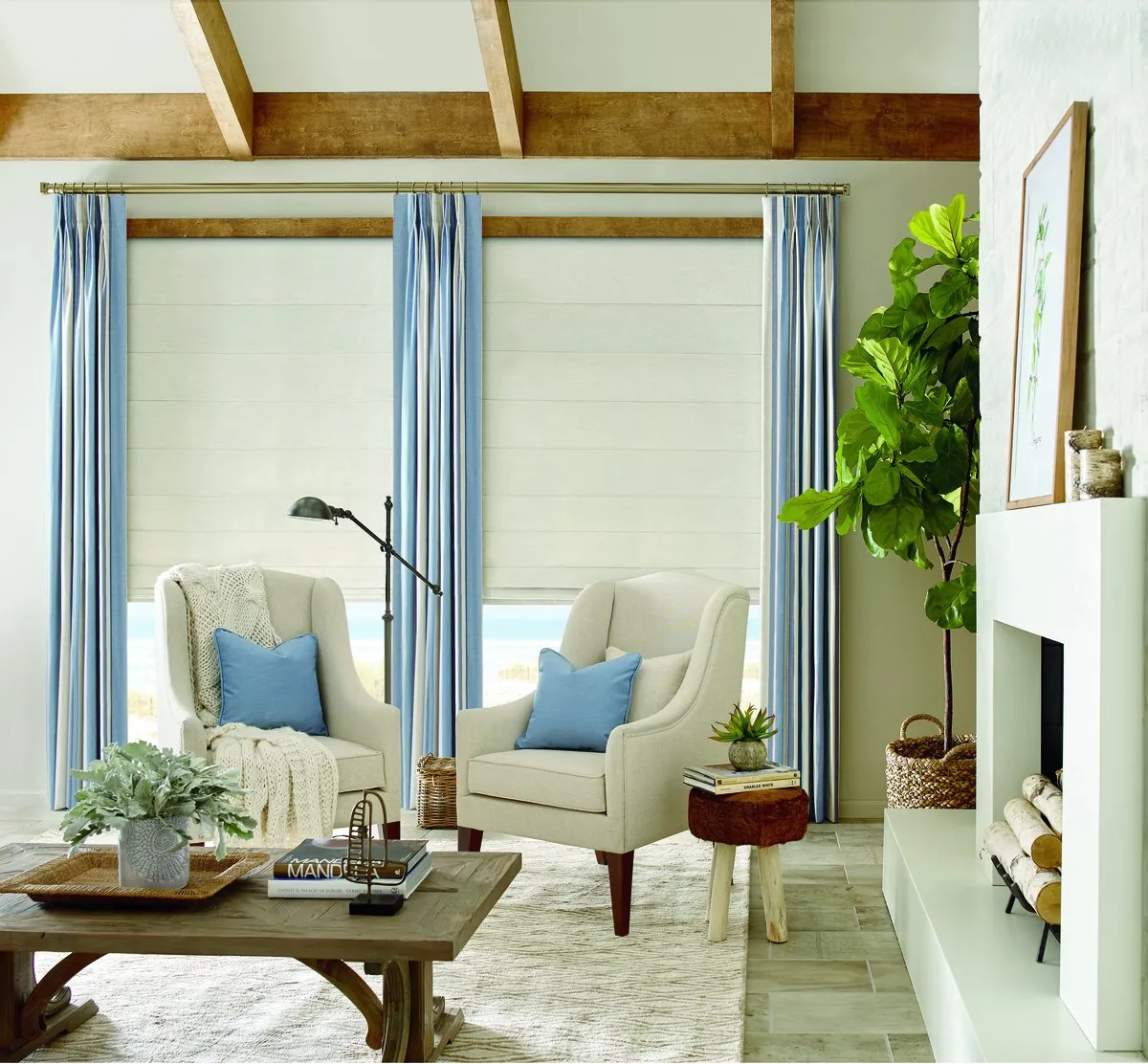 Cream roman shades on tall windows framed by blue striped drapery panels, in a living room with two armchairs, wood ceiling beams and a white fireplace