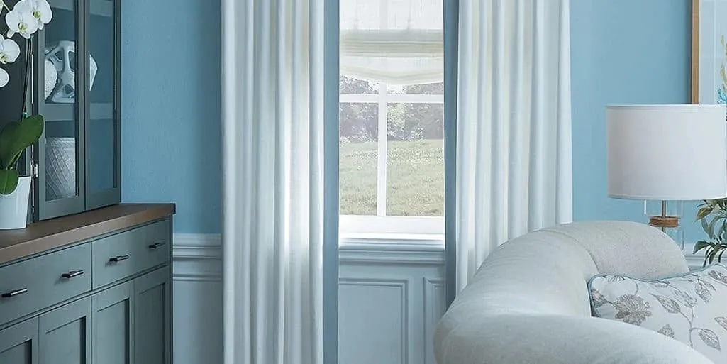 Floor-length white drapery panels framing a window in a blue living room, with a relaxed sheer shade behind