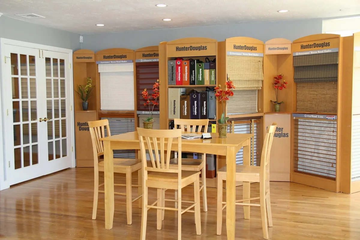 Showroom with a light wood table and four chairs in front of Hunter Douglas displays of woven wood shades, wood blinds and shelves of sample books