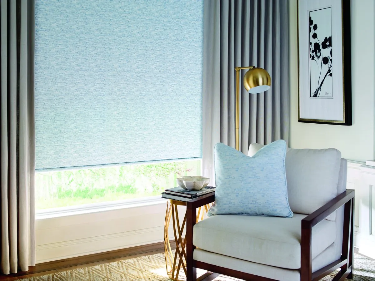Pale blue patterned roller shade lowered most of the way on a large window between grey drapery panels, beside a white armchair and brass floor lamp