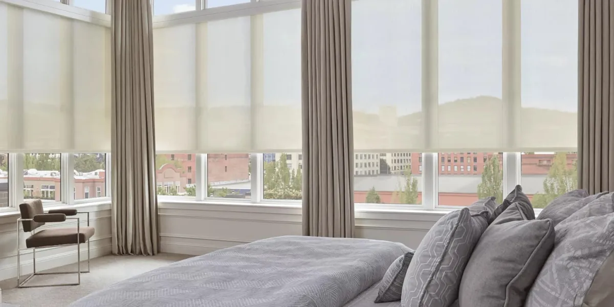 Ivory solar screen shades lowered partway on tall bedroom windows with taupe floor-length drapery and a city view