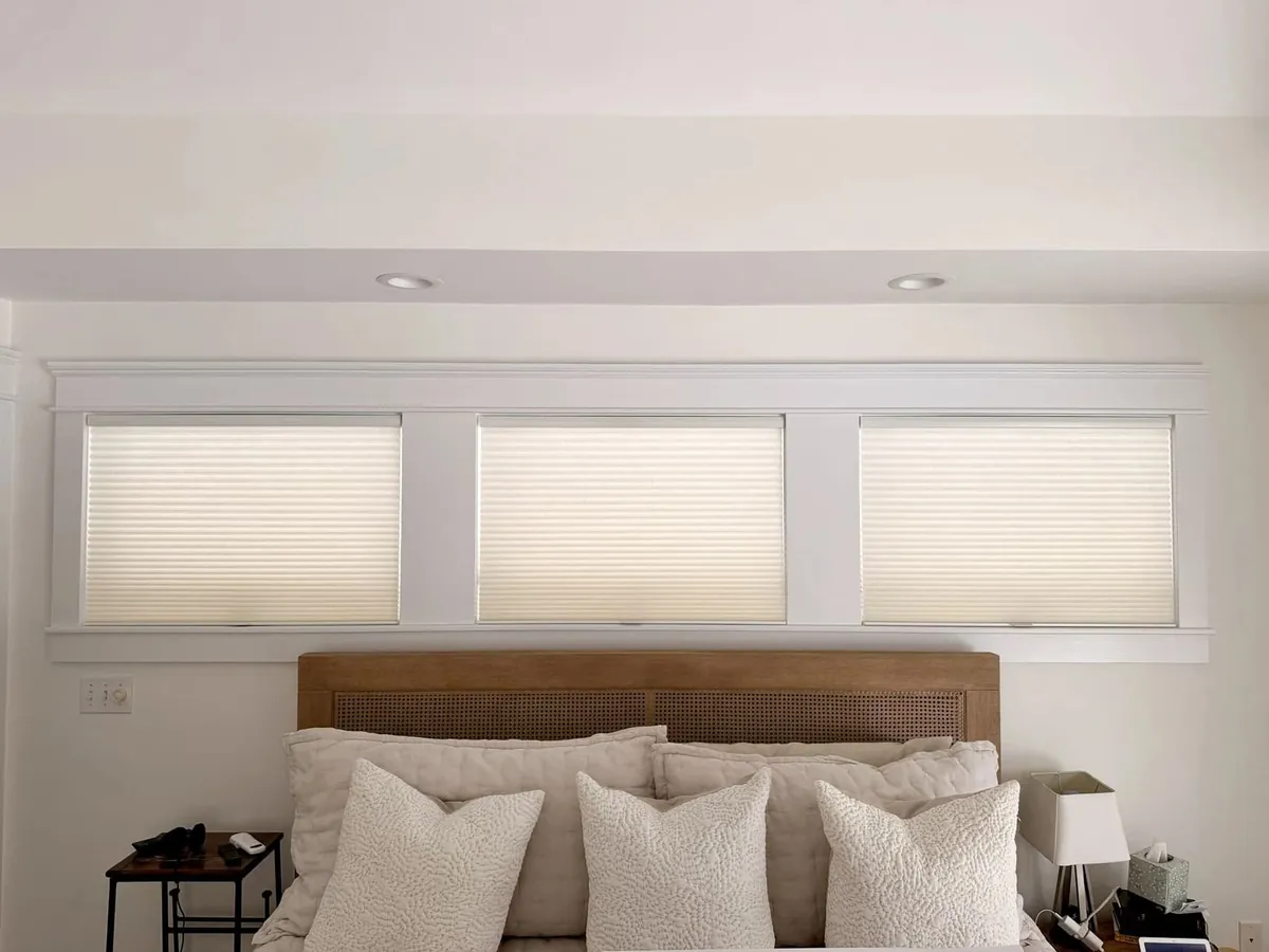 Three ivory honeycomb shades on a row of white-trimmed windows above a bed with a cane headboard and white pillows