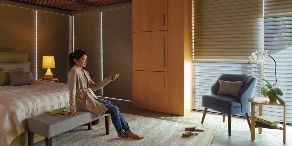 A woman seated on a bench holds a remote by sheer horizontal shades in a warm-toned bedroom