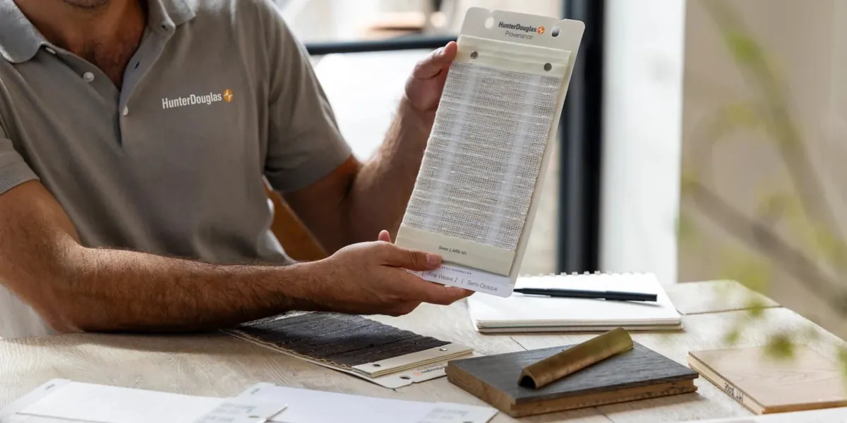 A consultant in a grey polo holds up a natural woven shade sample over a table of material swatches and a notebook