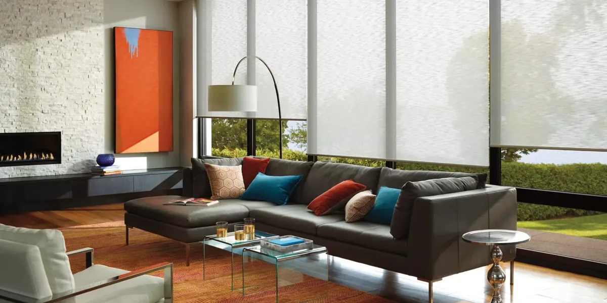 Light grey sheer roller shades partly lowered on a wall of windows in a modern living room with a dark leather sectional and fireplace