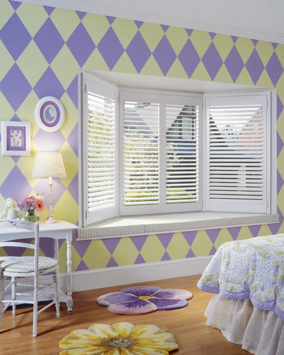 White plantation shutters on a bay window above a window seat in a child's bedroom with purple and yellow diamond walls