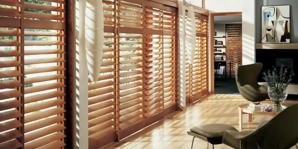 Stained wood shutters with louvres tilted open across a wall of windows, casting striped sunlight over a modern living room