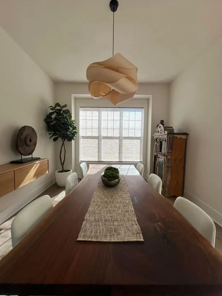 A natural woven shade lowered on the dining room window at the end of a long wood table under a pendant lamp