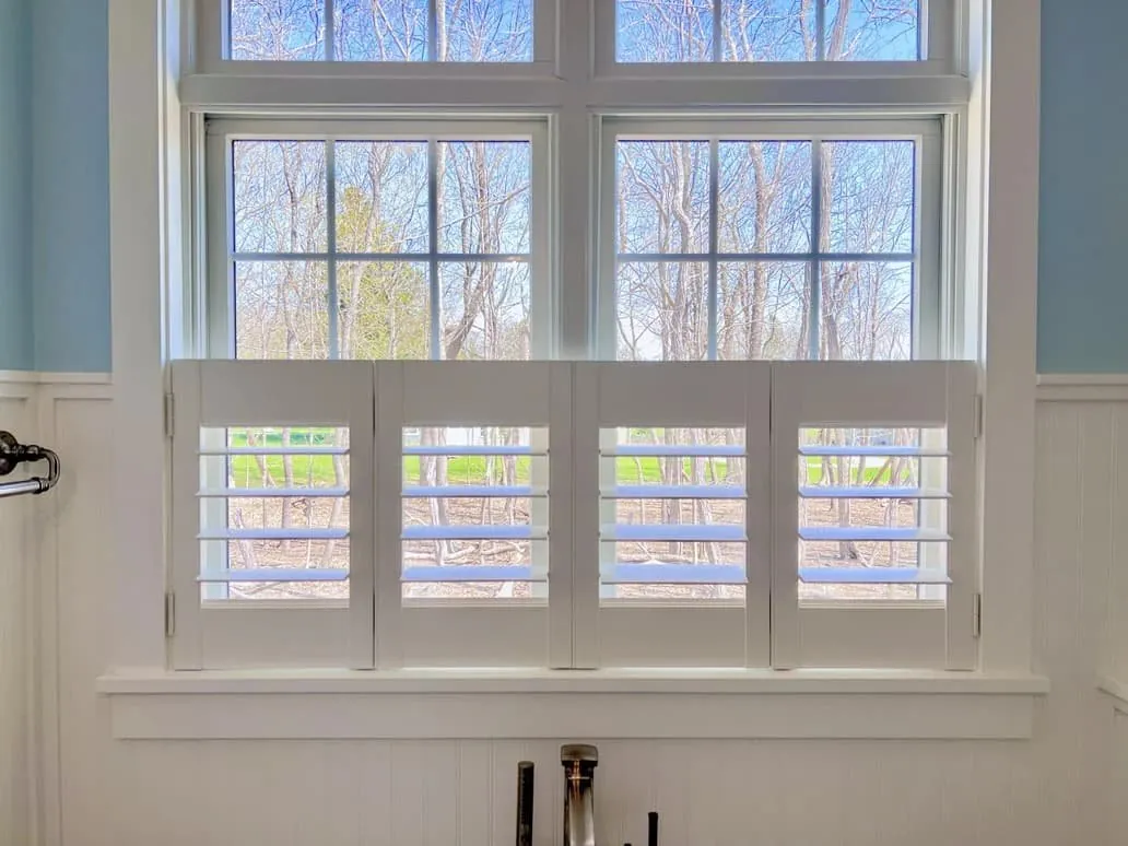 White cafe shutters with open louvres on the lower half of a double window above a sink, trees outside