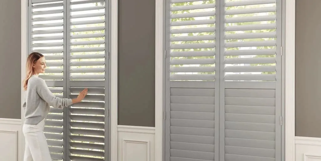 A woman opens grey painted shutters on tall windows, one panel's louvres tilted open to greenery outside