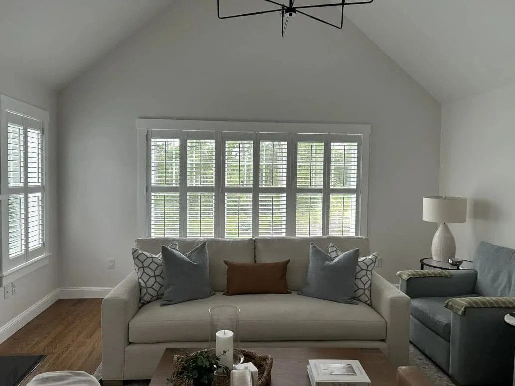 White plantation shutters on a wide window behind a cream sofa in a vaulted living room, plus a side window