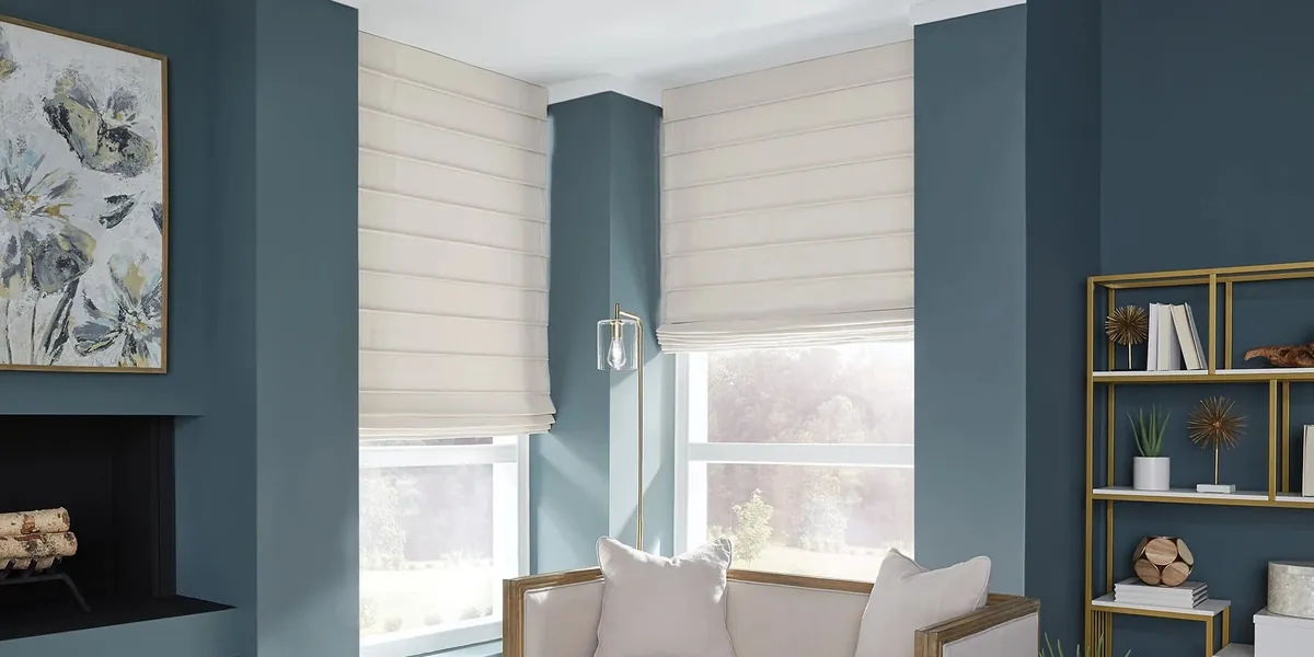 Cream flat-fold roman shades on bay windows in a living room with deep blue walls, a fireplace and gold shelving