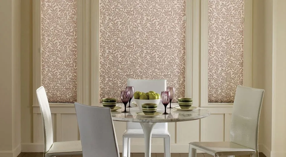Leaf-patterned fabric shades on three tall dining room windows behind a round marble table set with green bowls and purple glasses