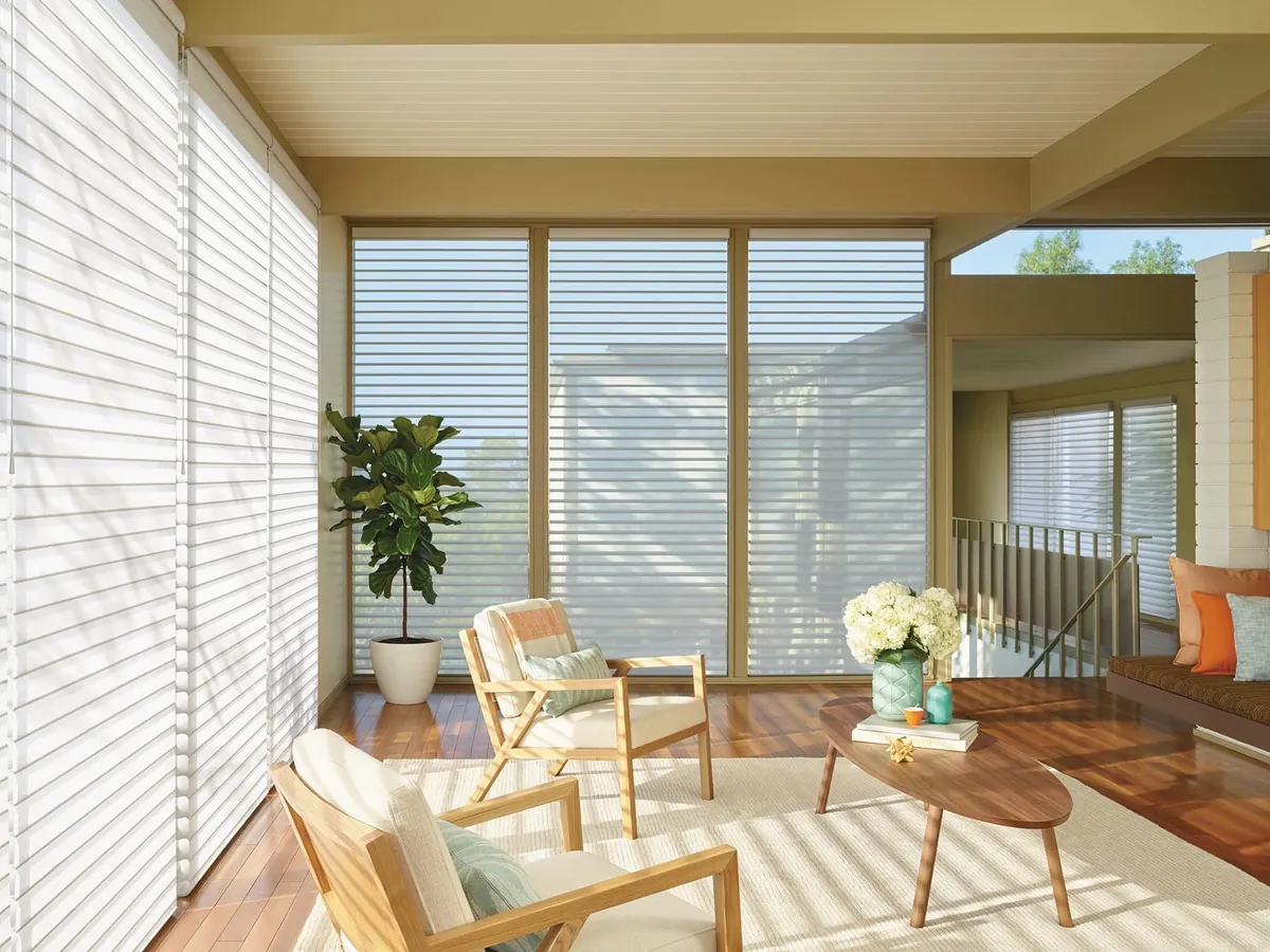 White sheer shades with horizontal fabric vanes on floor-to-ceiling windows in a sunny sitting room with two wood armchairs and an oval coffee table