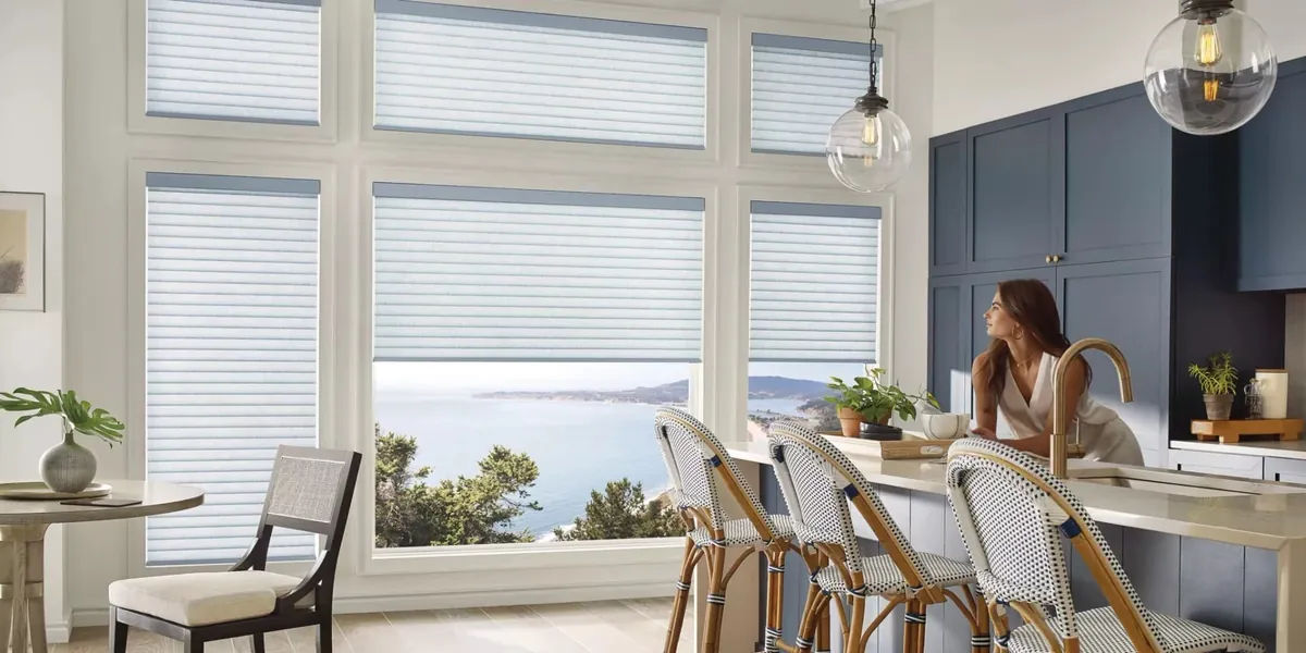 Soft white rolling shades with slate-blue headrails on tall kitchen windows overlooking the ocean, a woman leaning on the island