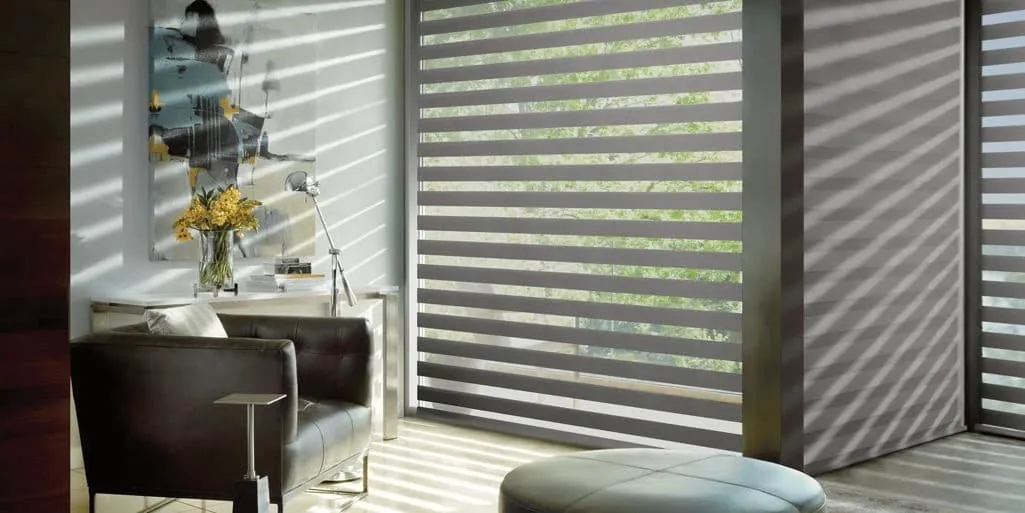 Grey banded shades with alternating sheer and solid stripes on large windows beside a leather armchair and ottoman