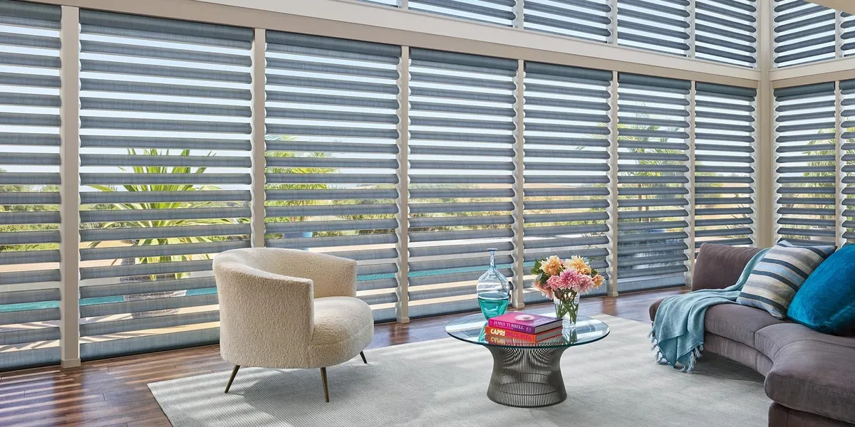 Blue-grey sheer shades with soft rounded vanes on a tall window wall in a living room with a cream armchair and glass table