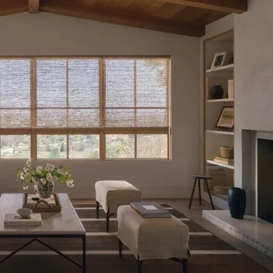 Natural woven shades on a wide window in a vaulted living room, with linen ottomans, a coffee table and a stone fireplace