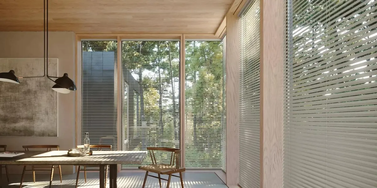 Thin silver aluminium blinds on floor-to-ceiling windows in a wood-lined dining room looking out to trees
