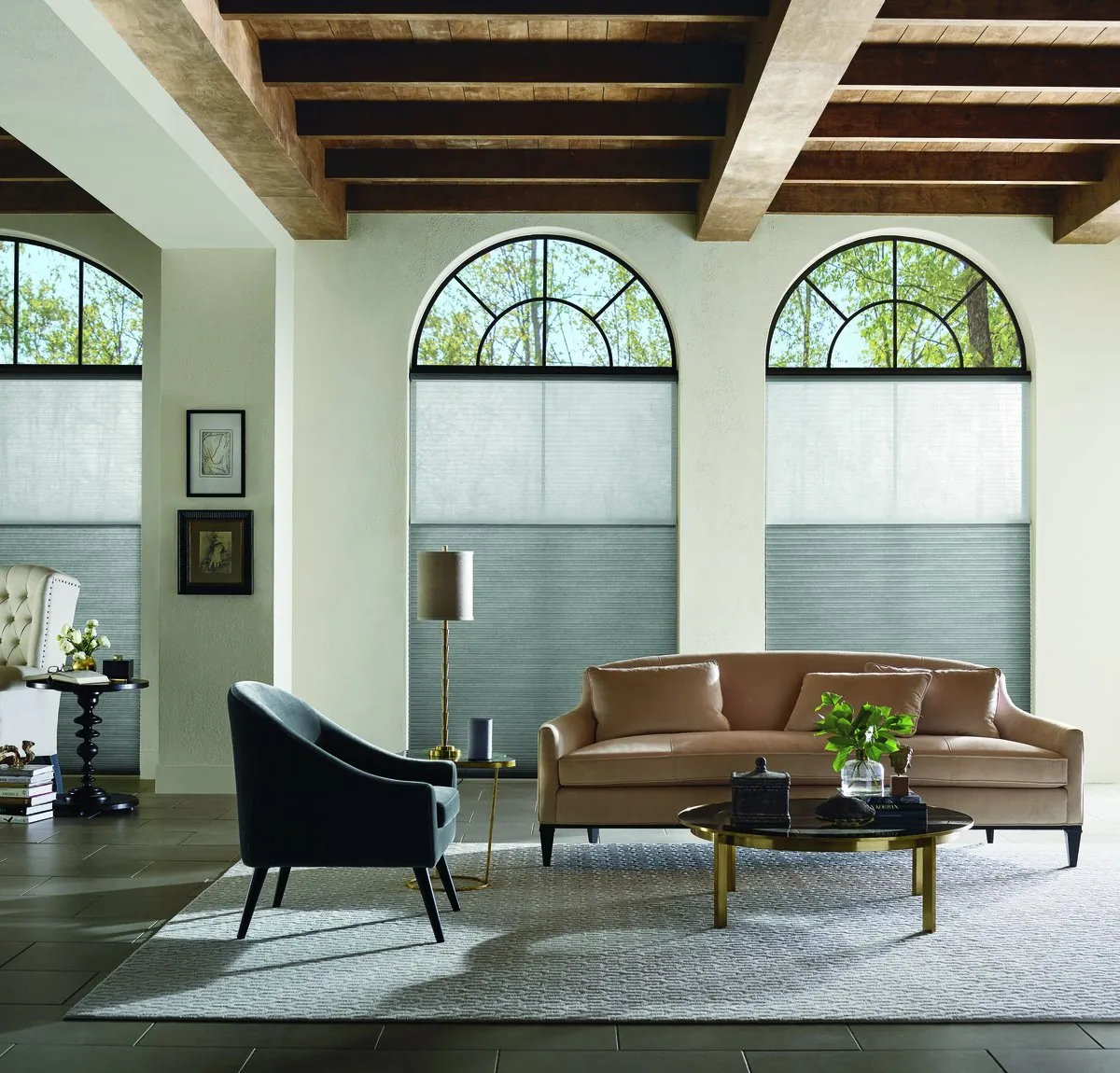 Grey honeycomb shades lowered from the top on tall arched windows, leaving the arches open, in a living room with wood ceiling beams and a tan sofa