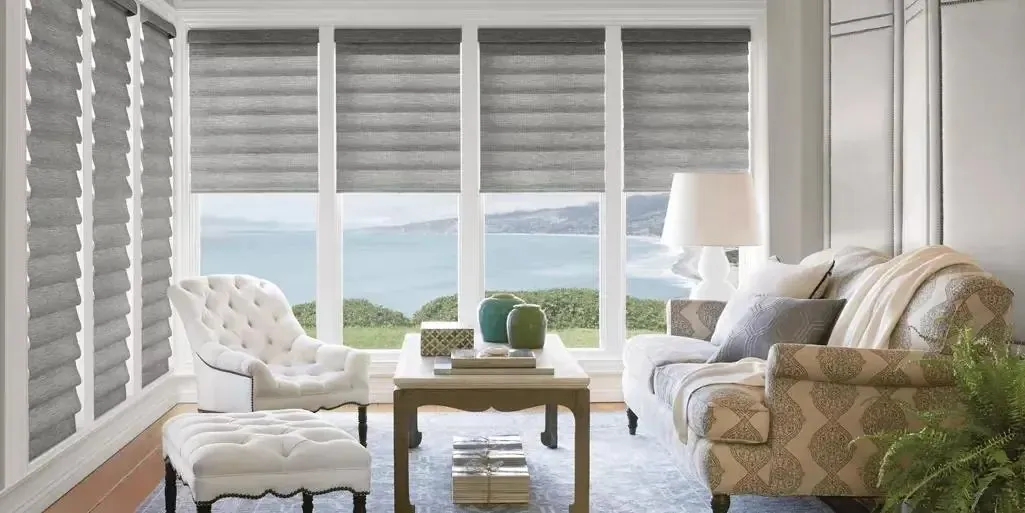 Grey roman shades with soft rounded folds across a sunroom of ocean-view windows, with a tufted white chair and patterned sofa